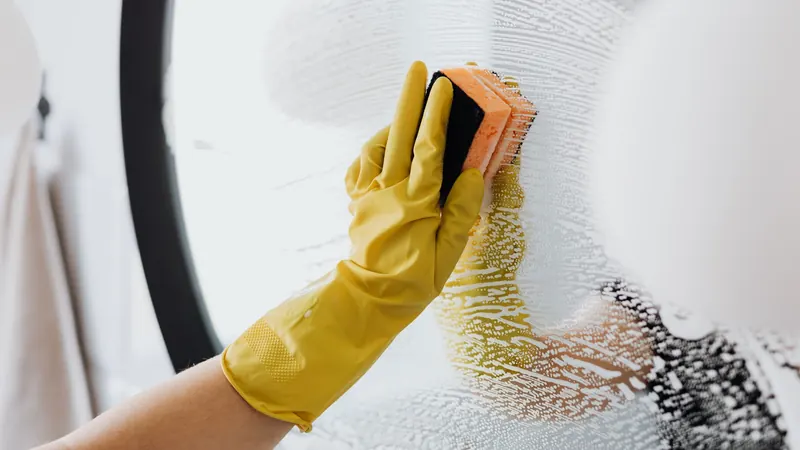 Gloved hand scrubbing a soapy bathroom mirror with a sponge during a timed house cleaning