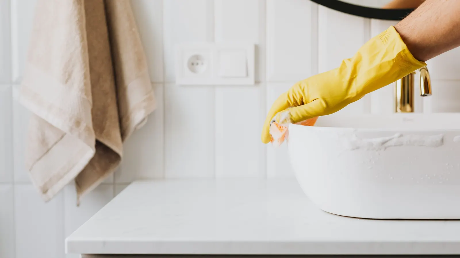 Hand in a yellow rubber glove wiping a white vessel sink in a tiled bathroom