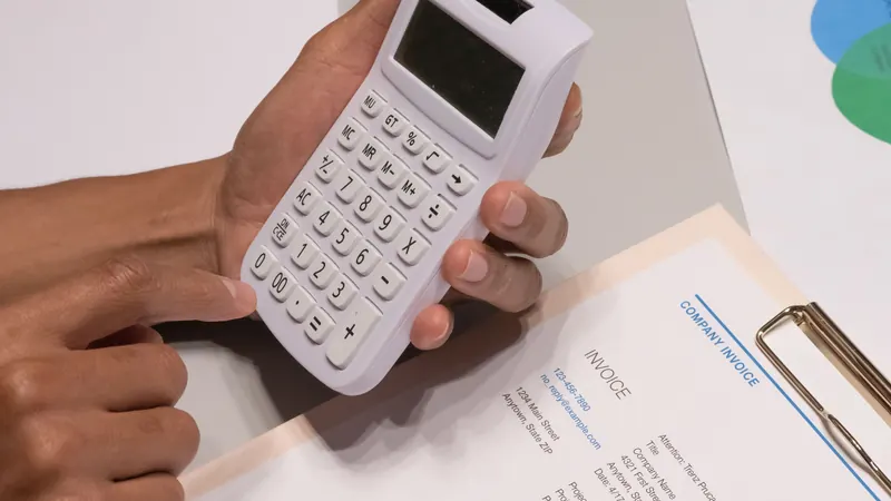 Hands holding a white calculator over a printed invoice on a clipboard