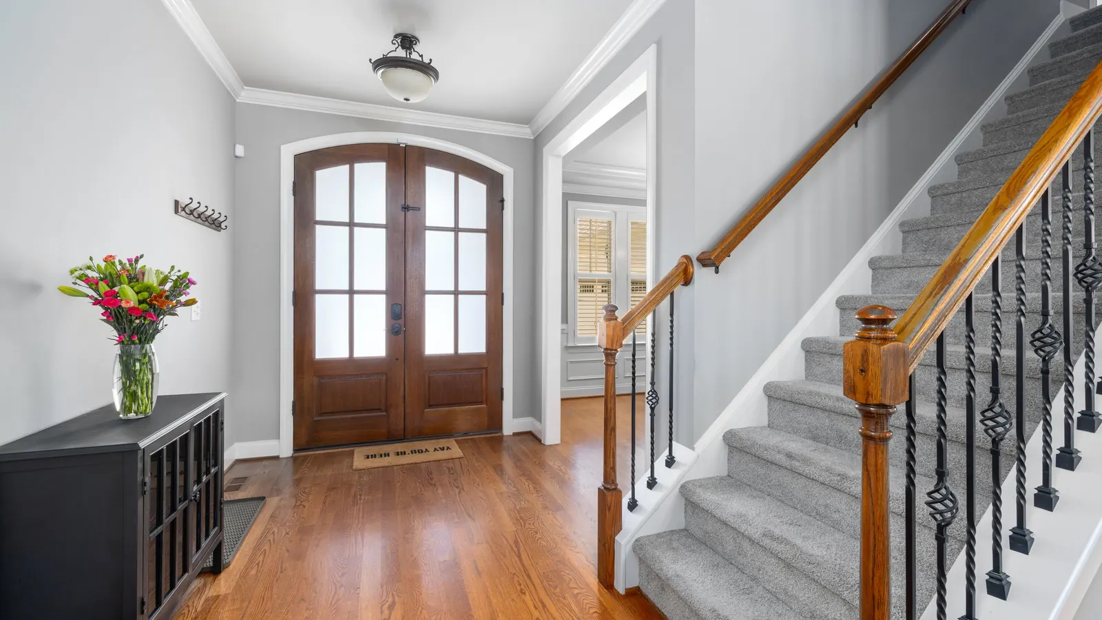 Carpeted staircase with wood handrail and iron balusters next to a front entry hall
