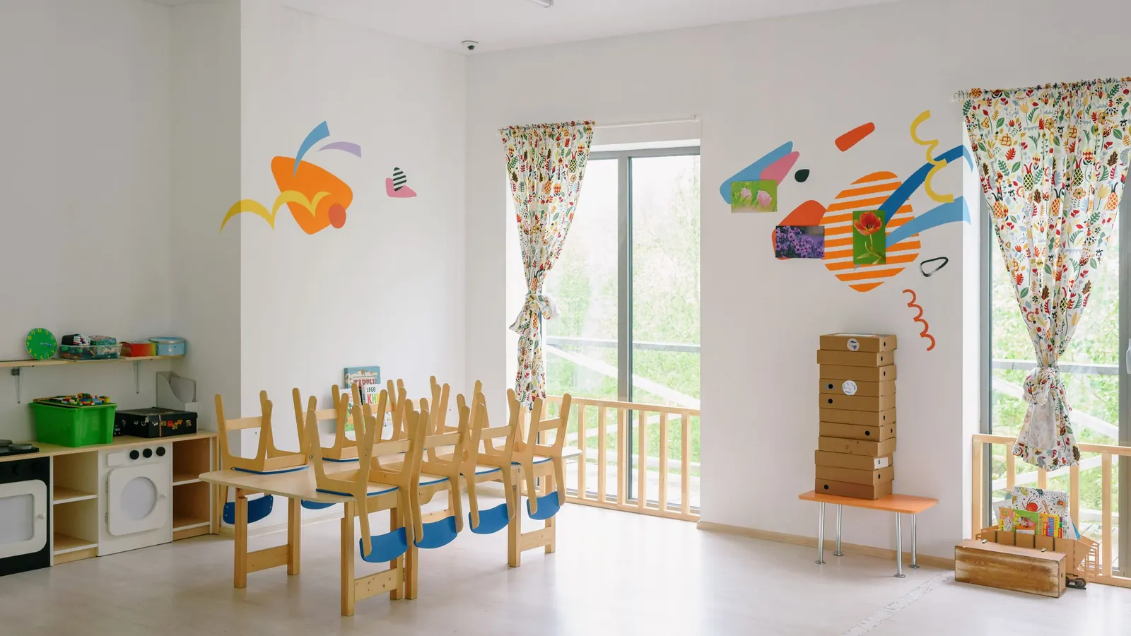 Empty daycare room with small chairs stacked on tables, toy kitchen and light wood floor