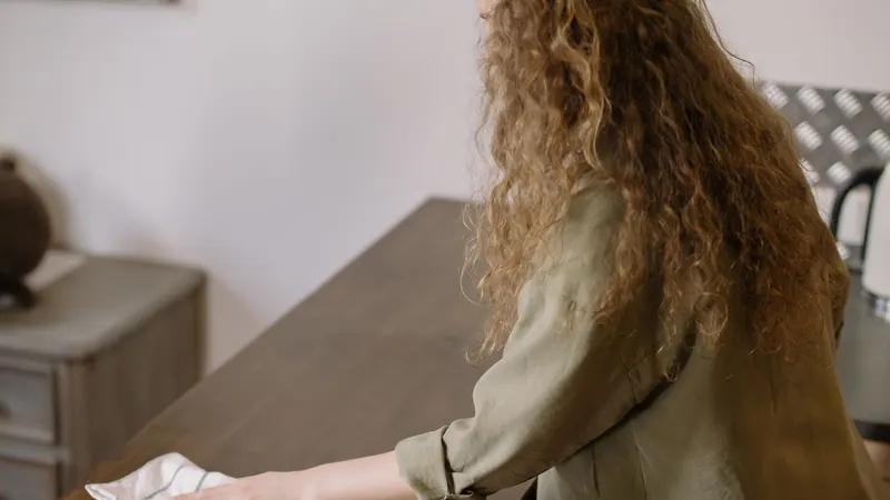 Hand wiping a wooden dresser top with a cloth, person shown from behind