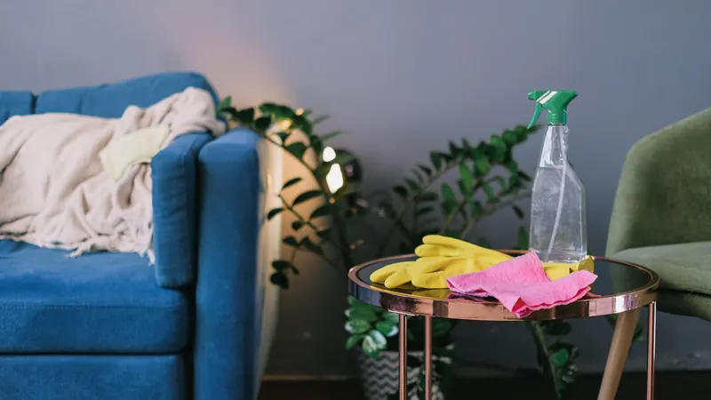 Yellow rubber gloves, pink cloth and a spray bottle on a side table next to a blue sofa