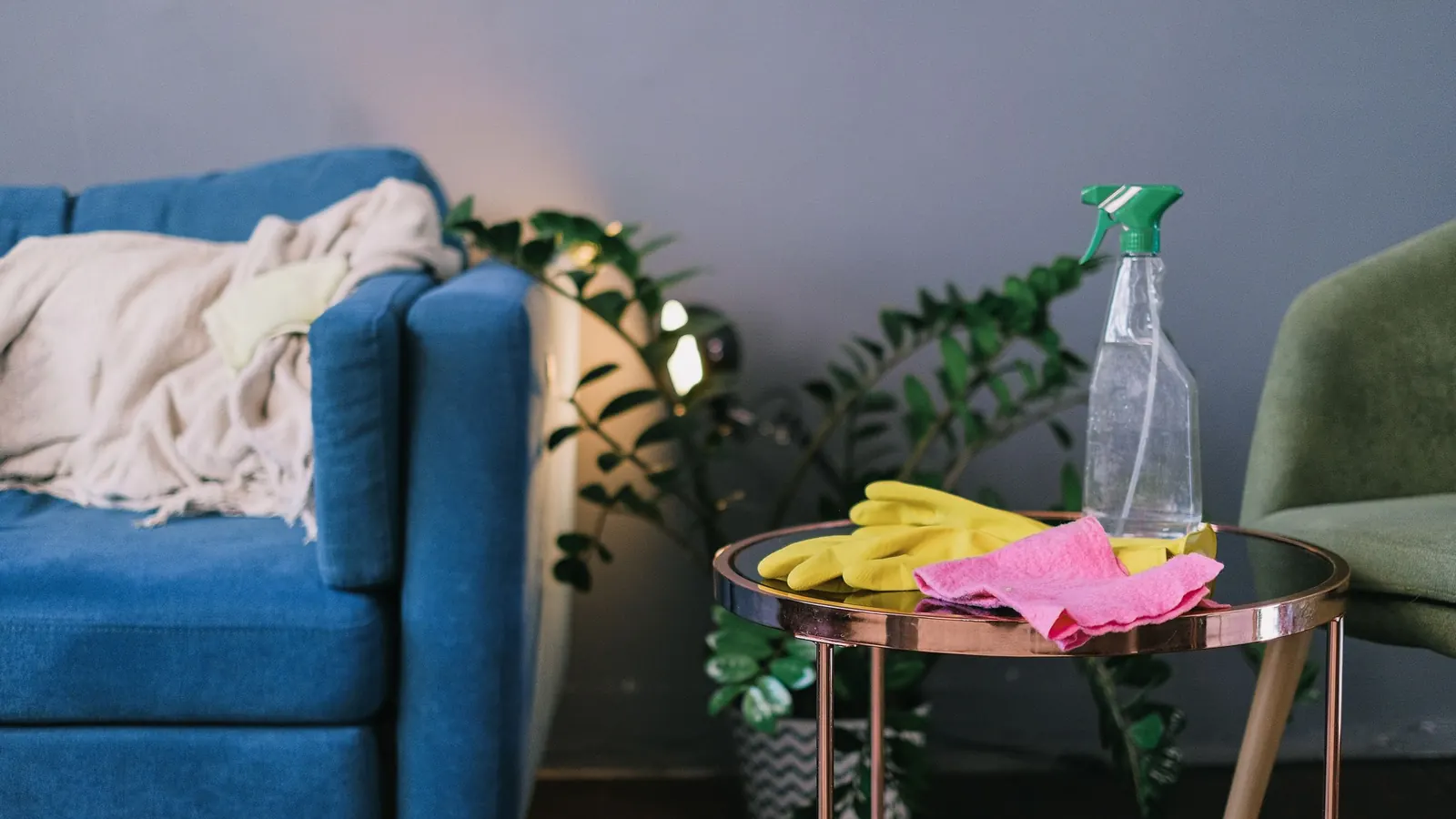 Yellow rubber gloves, pink cloth and a spray bottle on a side table next to a blue sofa