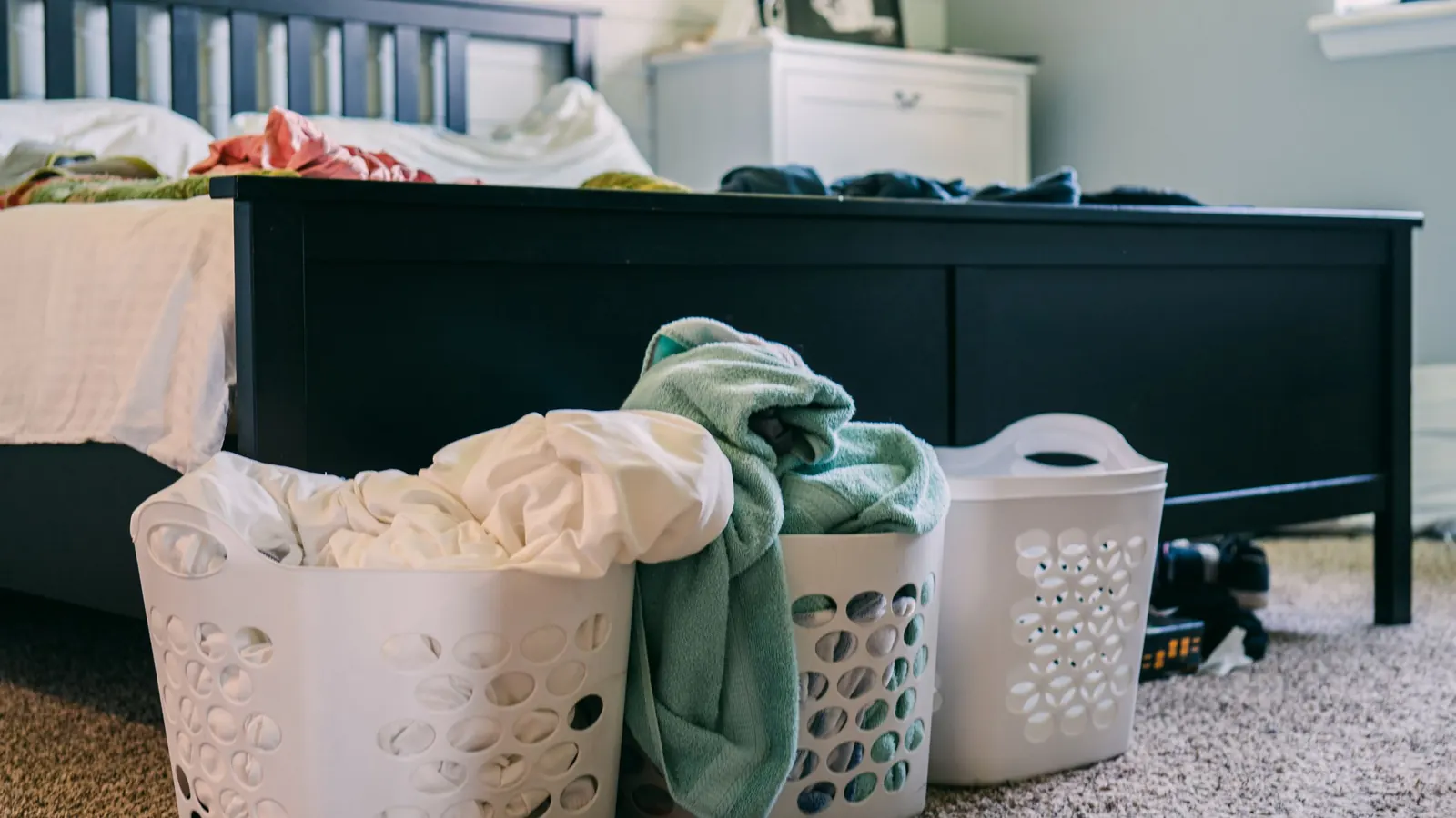 Laundry baskets full of clothes on a carpeted bedroom floor beside the bed, the pile that grows in the newborn weeks