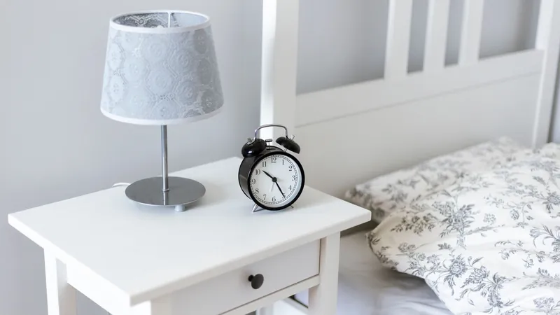 Bedside table with an alarm clock and lamp next to a made bed