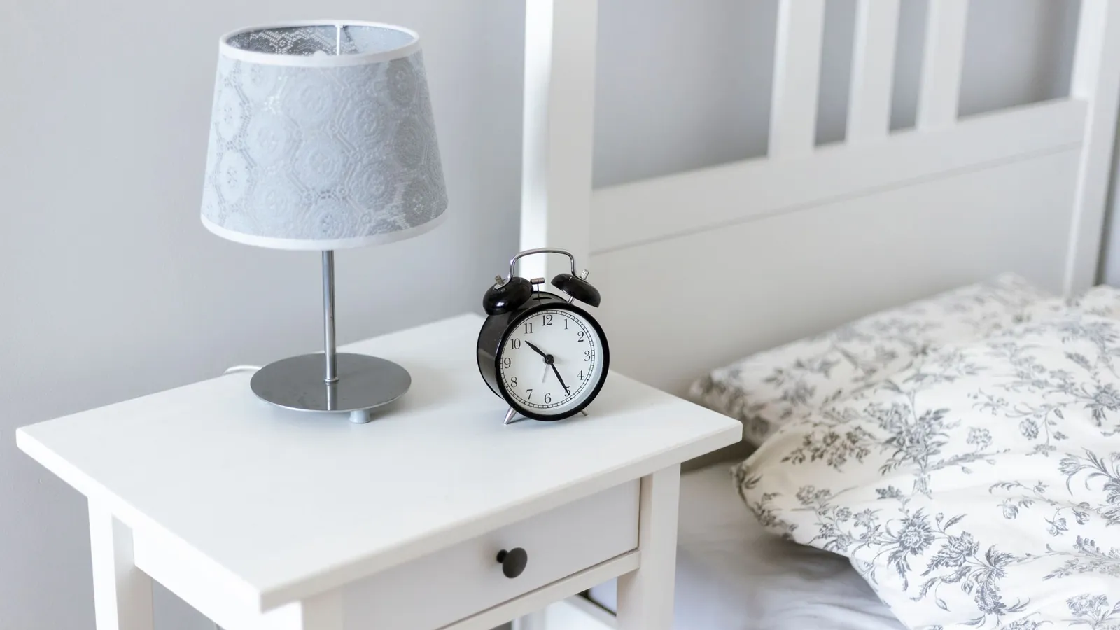 Bedside table with an alarm clock and lamp next to a made bed
