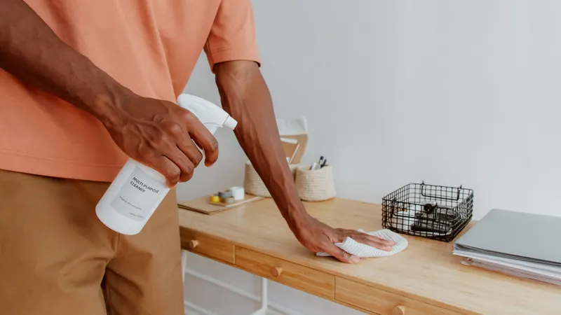 Person wiping a wooden desk with a cloth and spray bottle, face not shown