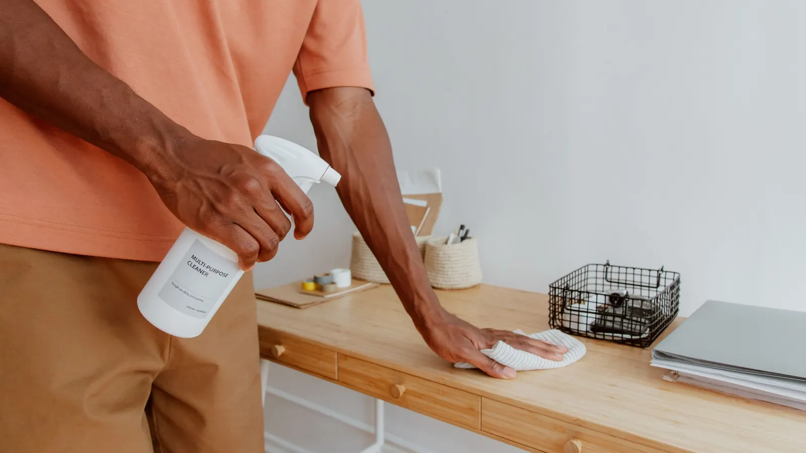 Person wiping a wooden desk with a cloth and spray bottle, face not shown